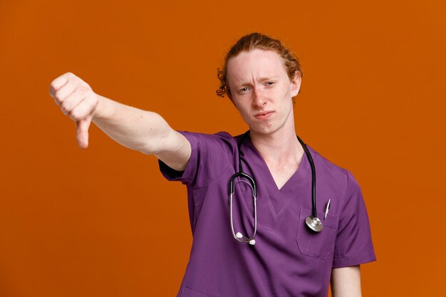 Desagradado mostrando os polegares para baixo jovem médico masculino vestindo uniforme com estetoscópio isolado em fundo laranja