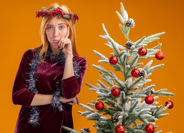 desagradável jovem linda em pé perto da árvore de natal usando vestido vermelho e grinalda com guirlanda no pescoço colocando a mão na bochecha isolada na parede laranja