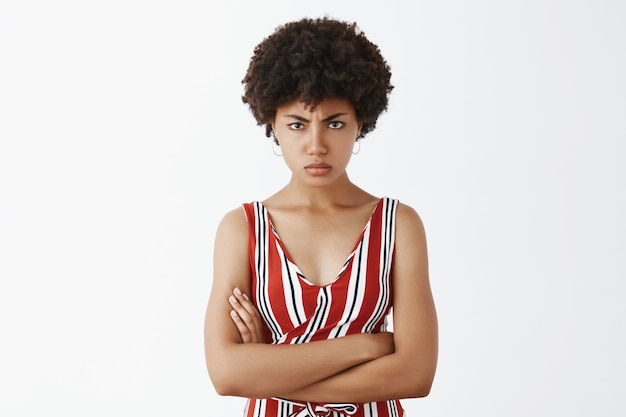 Foto grátis descontente, brava e irritada, atraente mulher afro-americana mandona de macacão listrado de mãos cruzadas no peito, carrancuda e amuada, decepcionada e ofendida