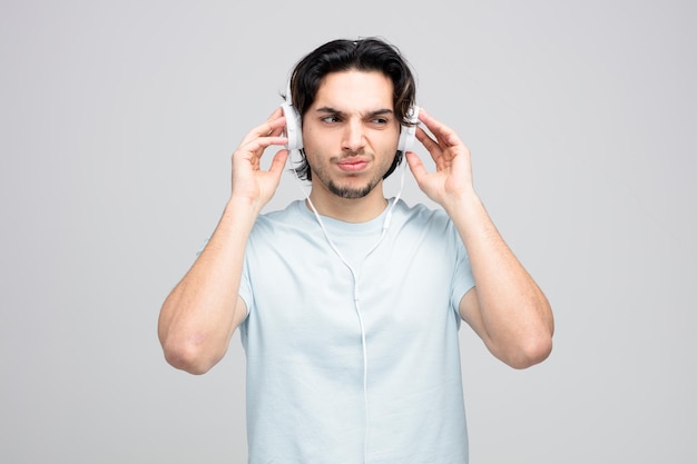 Descontente jovem bonito usando e tocando fones de ouvido olhando para o lado isolado no fundo branco