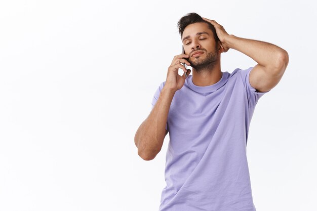 Descontraído e despreocupado, gay sonhador em camiseta roxa, penteando o cabelo com os dedos aliviados, olhos fechados falando no smartphone, tendo conversa sensual com namorado, gosto de ouvir a voz dele