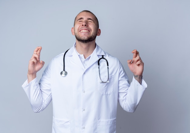Desejando um jovem médico vestindo túnica médica e estetoscópio ao redor do pescoço, fazendo gesto de dedos cruzados com os olhos fechados, isolado no fundo branco com espaço de cópia