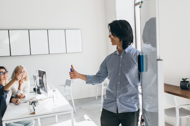 Desenvolvedor asiático em pé perto do flipchart e olhando para uma mulher loira de óculos. Retrato interior do empresário morena escrevendo gráfico no quadro branco e ouvindo colegas.