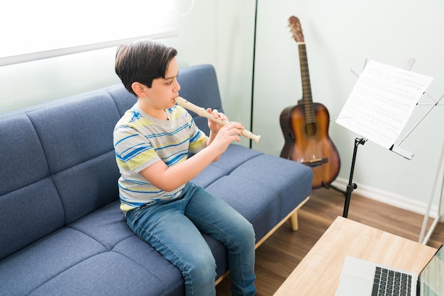 Foto grátis desenvolvendo minhas habilidades musicais. menino elementar caucasiano tocando sua música favorita na flauta enquanto está sentado na sala de estar para suas aulas on-line