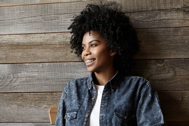 Desfrutando de um estilo de vida feliz e livre. Foto interior de mulher despreocupada bonita de pele escura relaxada com corte de cabelo Afro e aparelho sorrindo amplamente, tendo inspirado expressão sonhadora, relaxante na cafeteria