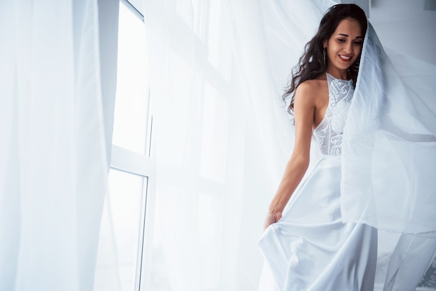 Desgaste elegante. Mulher bonita de vestido branco fica na sala branca com luz do dia através das janelas