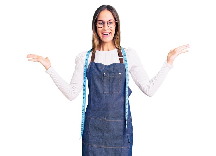Foto grátis designer de costureira linda morena jovem vestindo avental de atelier comemorando a vitória com sorriso feliz e expressão vencedora com as mãos levantadas