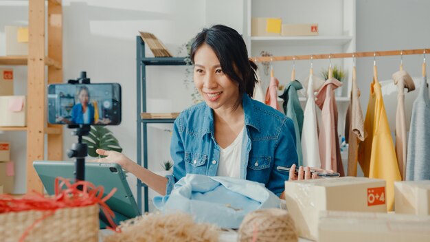 Designer de moda jovem asiática usando telefone celular, recebendo pedido de compra e mostrando roupas em transmissão ao vivo