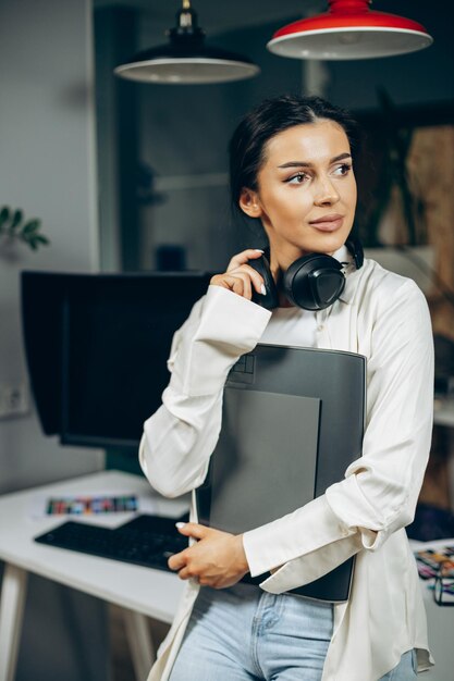 Designer de mulher com fones de ouvido musicais em pé no escritório segurando pasta