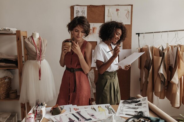 Designers de moda calmos e atraentes trabalham em um escritório aconchegante Tinta de mulher de pele escura Mulher de cabelo curto segura xícara de café