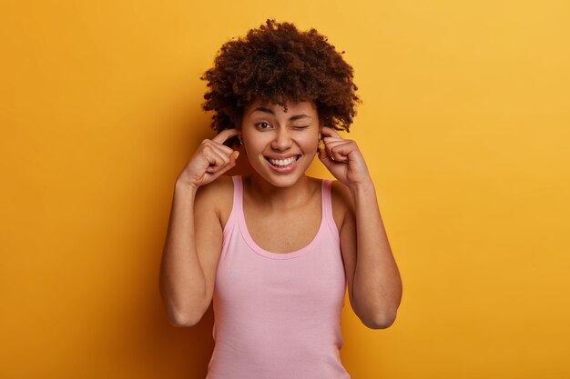 Desligue a música alta. Mulher afro-americana positiva tapa os ouvidos e pisca os olhos, sorri maliciosamente por causa do barulho, mostra os dentes brancos, ouve algo muito alto, vestida casualmente, posa contra a parede amarela