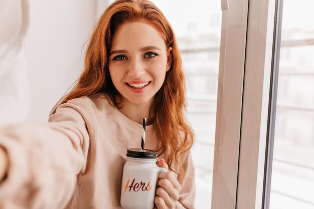 Deslumbrante mulher europeia bebendo cappuccino em casa Foto interior de uma linda garota ruiva fazendo selfie em seu quarto