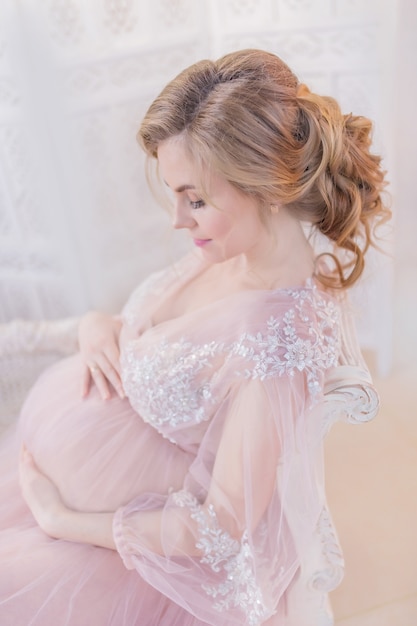 Deslumbrante mulher grávida no vestido rosa repousa no sofá em um quarto branco
