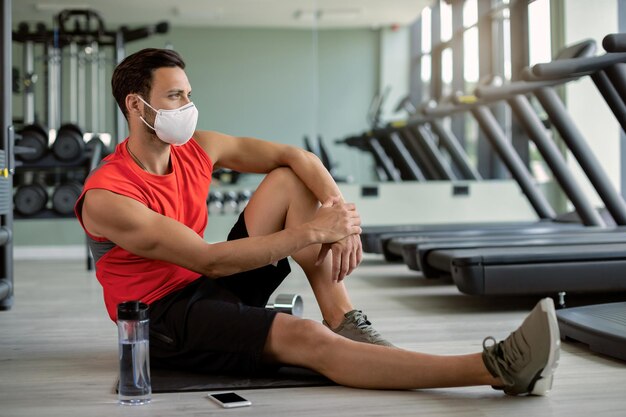 Desportista pensativo com máscara facial protetora relaxando no chão no health club