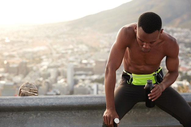 Desportivo cansado de pele escura, corre rápido na estrada da montanha, focado para baixo, recupera o fôlego, segura a garrafa de água doce, modelos contra a vista da montanha. espaço livre para seu conteúdo de publicidade