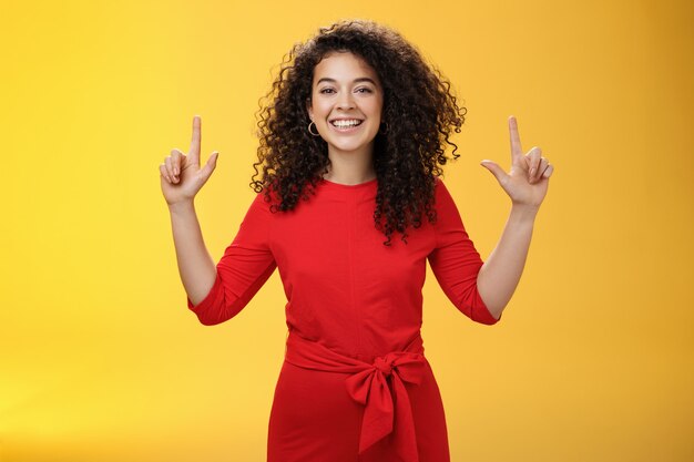 Despreocupada, alegre e amigável mulher de sucesso com penteado encaracolado em vestido vermelho sorrindo encantada e ambiciosa apontando para cima com as mãos levantadas dando recomendação ou mostrando promoção.