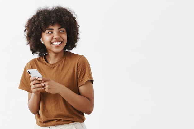 Despreocupada, amigável e criativa, elegante adolescente afro-americana com camiseta marrom, virando à direita com largo sorriso satisfeito, usando fones de ouvido sem fio e segurando um smartphone