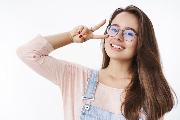 Foto grátis despreocupada atraente jovem carismática 20 anos mulher de óculos com cabelo castanho, mostrando vitória ou gesto de paz perto do rosto inclinando a cabeça com alegria e sorrindo satisfeito na frente posando sobre parede cinza.