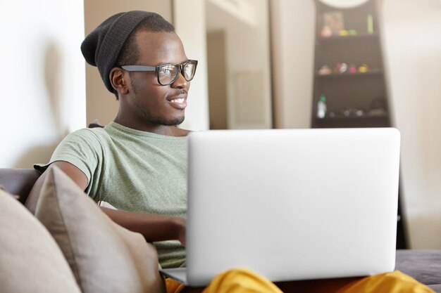 Despreocupada e descontraída jovem estudante de pele escura de chapéu e óculos relaxando em casa depois da faculdade, sentada no sofá com o laptop no colo, jogando videogame ou assistindo séries favoritas online