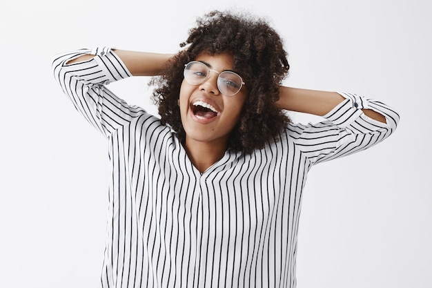 despreocupada, feliz e alegre, bonita, de pele escura, com penteado afro, de mãos dadas atrás da cabeça e sorrindo, satisfeita e relaxada levando um estilo de vida elevado