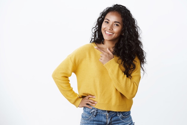 Despreocupada jovem afro-americana moderna com cabelo encaracolado em um suéter amarelo, incline a cabeça com alegria e sorria, observe a câmera enquanto discute uma nova promoção legal, apontando para o espaço em branco esquerdo sobre a parede branca