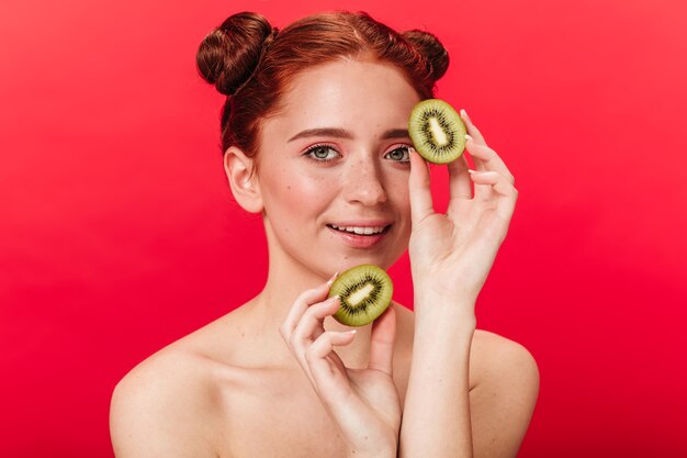 Despreocupada senhora de gengibre segurando kiwi. Foto de estúdio de elegante mulher caucasiana com frutas exóticas isoladas em fundo vermelho.