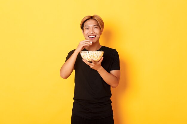 Despreocupado feliz asiático com cabelo loiro, assistindo comédia, rindo e comendo pipoca, em pé na parede amarela.
