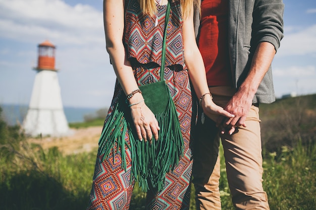 Detalhes de moda de mãos dadas com um casal jovem hippie em estilo indie apaixonado caminhando pelo campo, farol no fundo