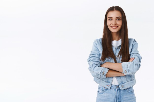 Determinada, confiante e feliz menina morena em jaqueta jeans, jeans, braços cruzados sobre o peito, sorrindo otimista, parecendo assertiva, autoconfiante, de pé despreocupada sobre um fundo branco com um sorriso amigável