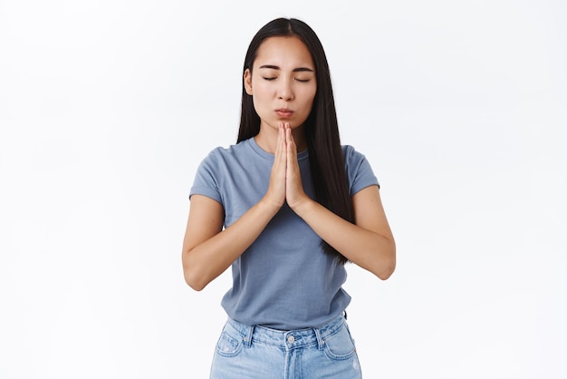 Determinada garota asiática bonita, séria e focada, rezando pelo bem-estar e o sonho se torna realidade em pé perto do santuário, feche os olhos, pedindo ajuda a deus, pressione as mãos juntas em gesto de oração