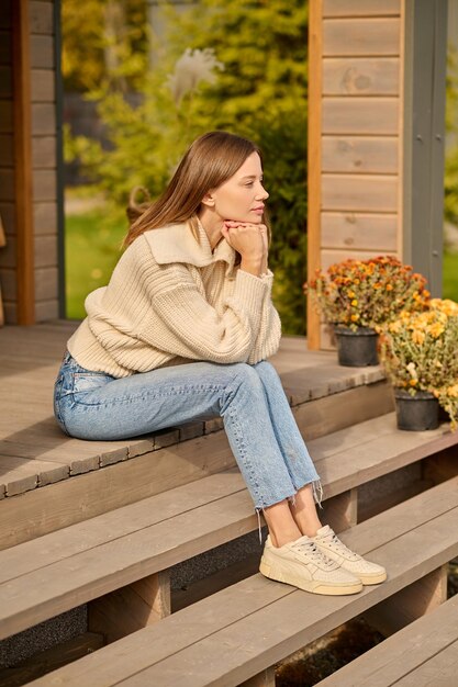 Dia bom. Jovem mulher bonita em roupas casuais tocando as mãos no queixo sentado pensativo na varanda da casa de campo lateralmente para a câmera no dia ensolarado de outono