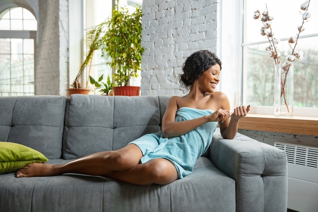 Dia da Beleza. Mulher afro-americana na toalha, preparada para fazer sua rotina diária de cuidados com a pele em casa. Sentado no sofá, fazendo as unhas, sorrindo. Conceito de beleza, autocuidado, cosméticos, juventude, saúde.