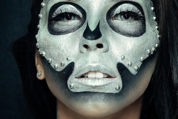 Dia das bruxas, feriados, estilo de vida, pessoas, beleza, conceito criativo - tema halloween e maquiagem criativa: modelo de menina bonita com corpo preto com máscara prateada pintura do crânio em fundo escuro no estúdio