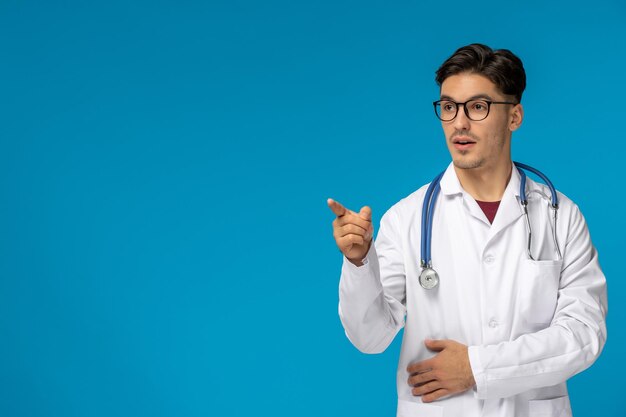 Dia dos médicos bonitão moreno bonitinho em vestido médico usando óculos e apontando para a esquerda