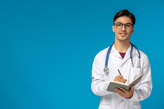 Dia dos médicos bonito jovem bonito de jaleco e óculos escrevendo no caderno