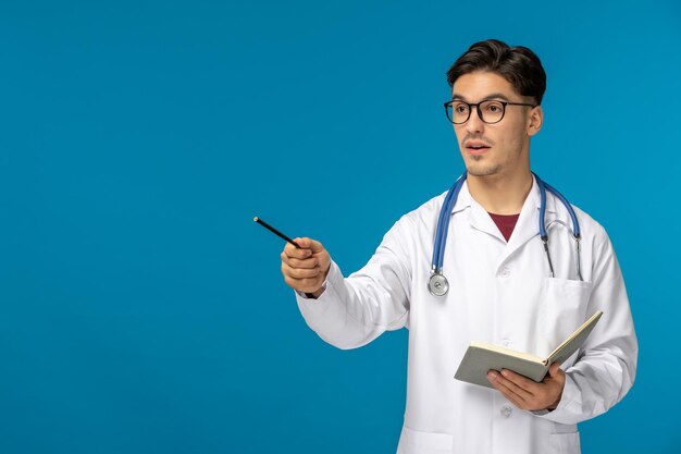 Dia dos médicos bonito jovem bonito de jaleco e óculos segurando caneta e caderno