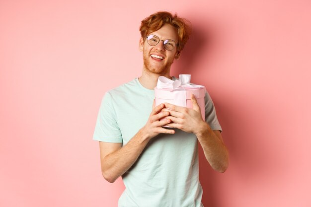 Dia dos namorados e conceito de romance. Feliz namorado ruivo recebe presente romântico, abraça a caixa com presente e diga obrigado, sorrindo agradecido, fundo rosa.
