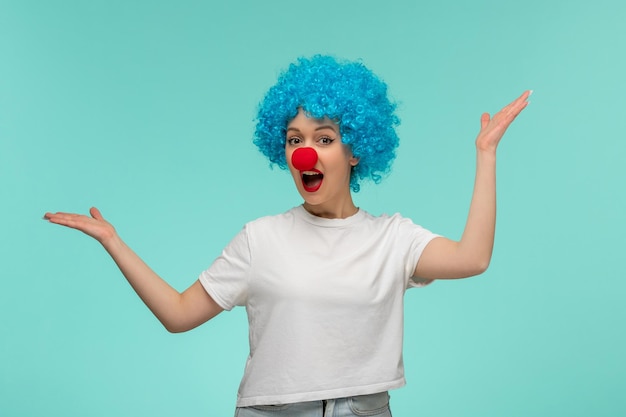 Dia dos tolos de abril, garota surpresa, acenando com as mãos, boca aberta com nariz vermelho em um traje de palhaço, cabelo azul