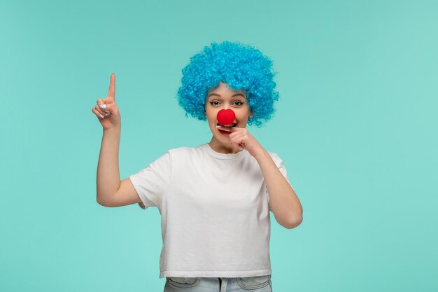 Dia dos tolos de abril linda garota apontando para cima mordendo o dedo com nariz vermelho em uma fantasia de palhaço cabelo azul