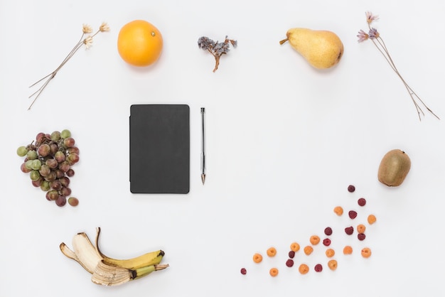 Diário de capa preta fechada e caneta rodeada com muitas frutas em pano de fundo branco