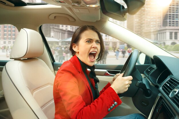 Dirigindo pela cidade. Jovem mulher atraente dirigindo um carro. Jovem modelo muito caucasiano no elegante elegante casaco vermelho sentado no interior do veículo moderno. Conceito de mulher de negócios.