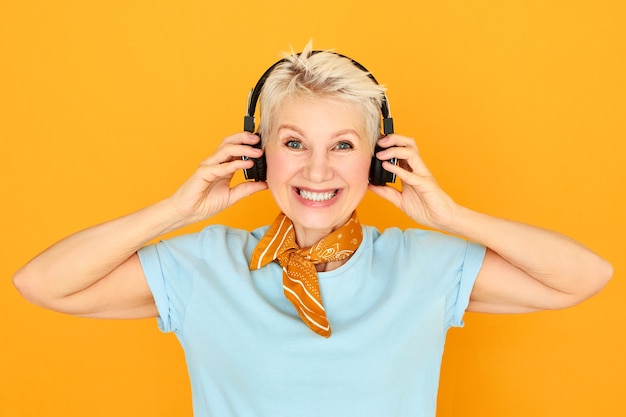 Foto grátis dispositivos eletrônicos, entretenimento, aposentadoria e conceito de idade. mulher loira feliz e encantadora aposentada usando fones de ouvido pretos sem fio, curtindo um som de áudio de alta resolução e ouvindo música