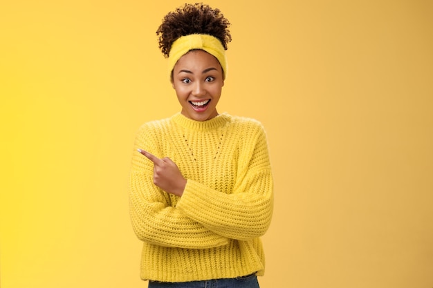 Divertida carismática sorridente linda garota negra no suéter com bandana arregalar os olhos caírem da mandíbula espantado ouvir sobre um novo lugar interessante e incrível amigo falando em pé fundo amarelo apontando para a esquerda espantado.