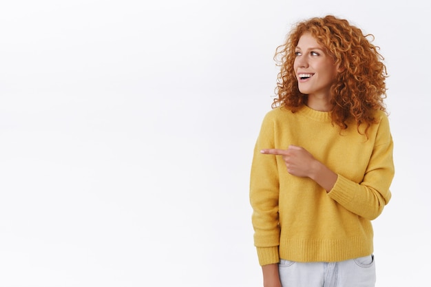 Divertida e atraente garota ruiva cacheada com suéter branco, comparecer à festa, ver amigos e apontar o dedo, virar à esquerda, sorrindo maravilhada e feliz, evento legal de check-out, parede branca