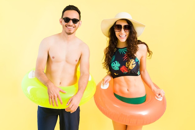 Divertido casal jovem em trajes de banho vestindo salva-vidas infláveis e sorrindo enquanto caminhava para a praia para entrar no mar