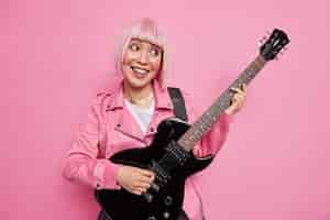 Foto grátis divertido e alegre estrela do rock de cabelo rosa toca guitarra elétrica sendo parte da banda vestida com uma jaqueta pronta para tocar no palco executa novas poses de música indoor e se diverte. conceito de passatempo de entretenimento musical