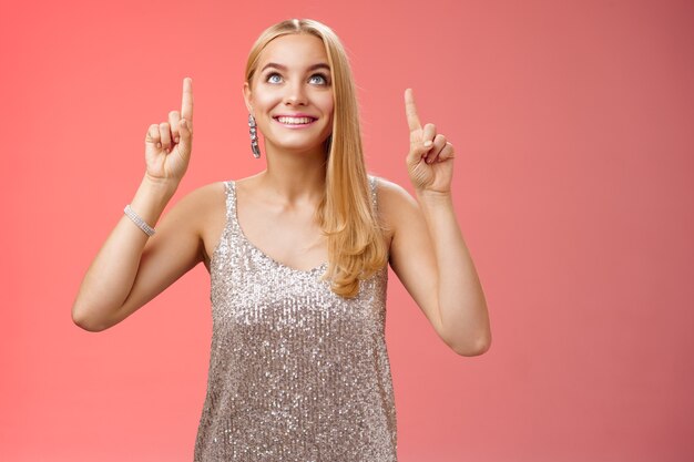 Divertido lindo concurso elegante mulher europeia loira longo penteado em prata brilhante vestido de noite olhar apontando para cima olhar curioso excitação ver a coisa desejada, em pé fundo vermelho alegre.