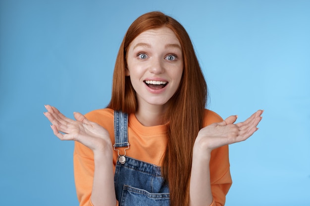 Divertido surpreso feliz jovem ruiva sem palavras feliz ver amigo voltar cidade natal olhos arregalados impressionados sorrindo levantar as mãos para os lados totalmente descrença em pé fundo azul alegre