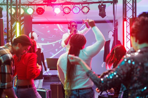 Dj falando para o público no microfone durante a apresentação no palco em boate. Pessoas dançando e levantando as mãos na pista de dança enquanto assistem a um concerto de música eletrônica no clube