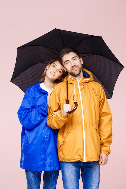 Doce casal lindo jovem posando em casacos de chuva segurando guarda-chuva sobre parede rosa clara